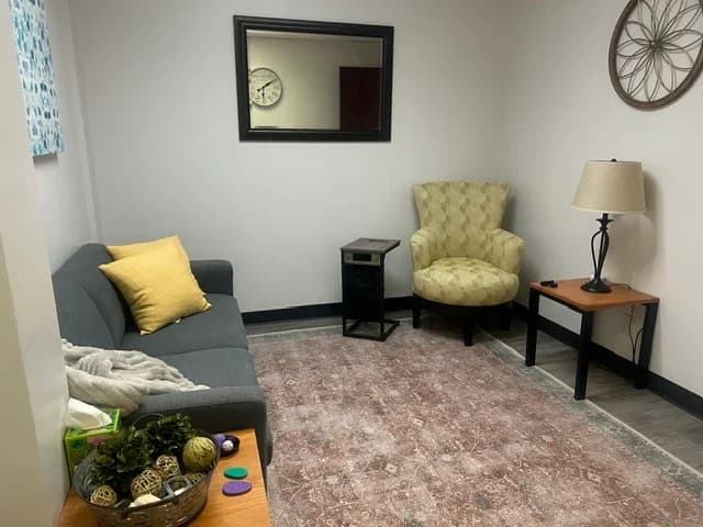 A therapy room with a grey sofa, a green upholstered armchair, and a table lamp beside a framed mirror