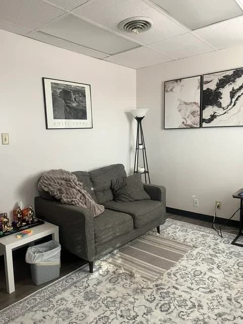 A quiet counseling room with a grey loveseat, a soft throw, framed prints, and a floor lamp in the corner