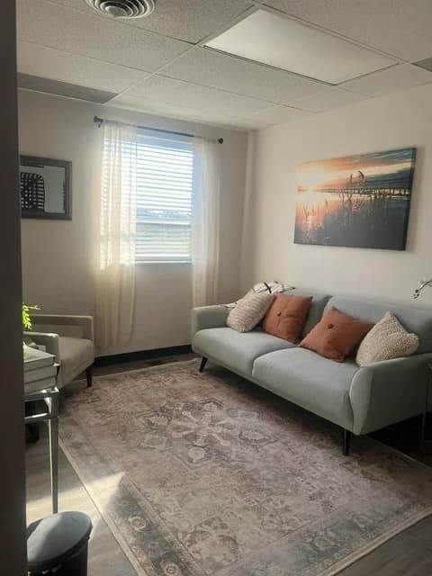 A therapy room with daylight through a curtained window, a pale sofa with cushions, and an armchair on a patterned rug